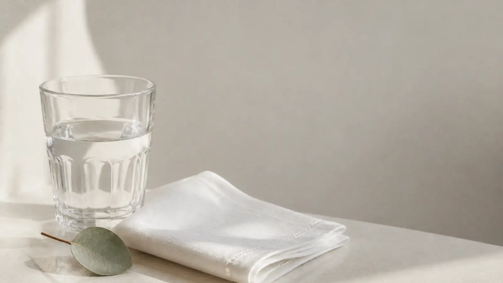 Gefaltetes weißes Leinentuch, ein Glas Wasser und ein Eukalyptusblatt auf hellem Grund